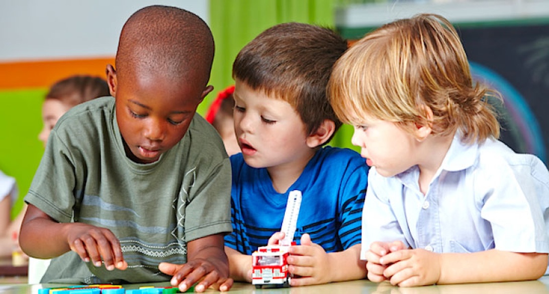 Three young children are gathered around a table in what appears to be an educational setting, deeply engaged in examining or working with a small toy or educational tool. The colorful background suggests a classroom environment designed for young learners. This image captures peer-based learning and hands-on education, important aspects of early childhood educational environments.